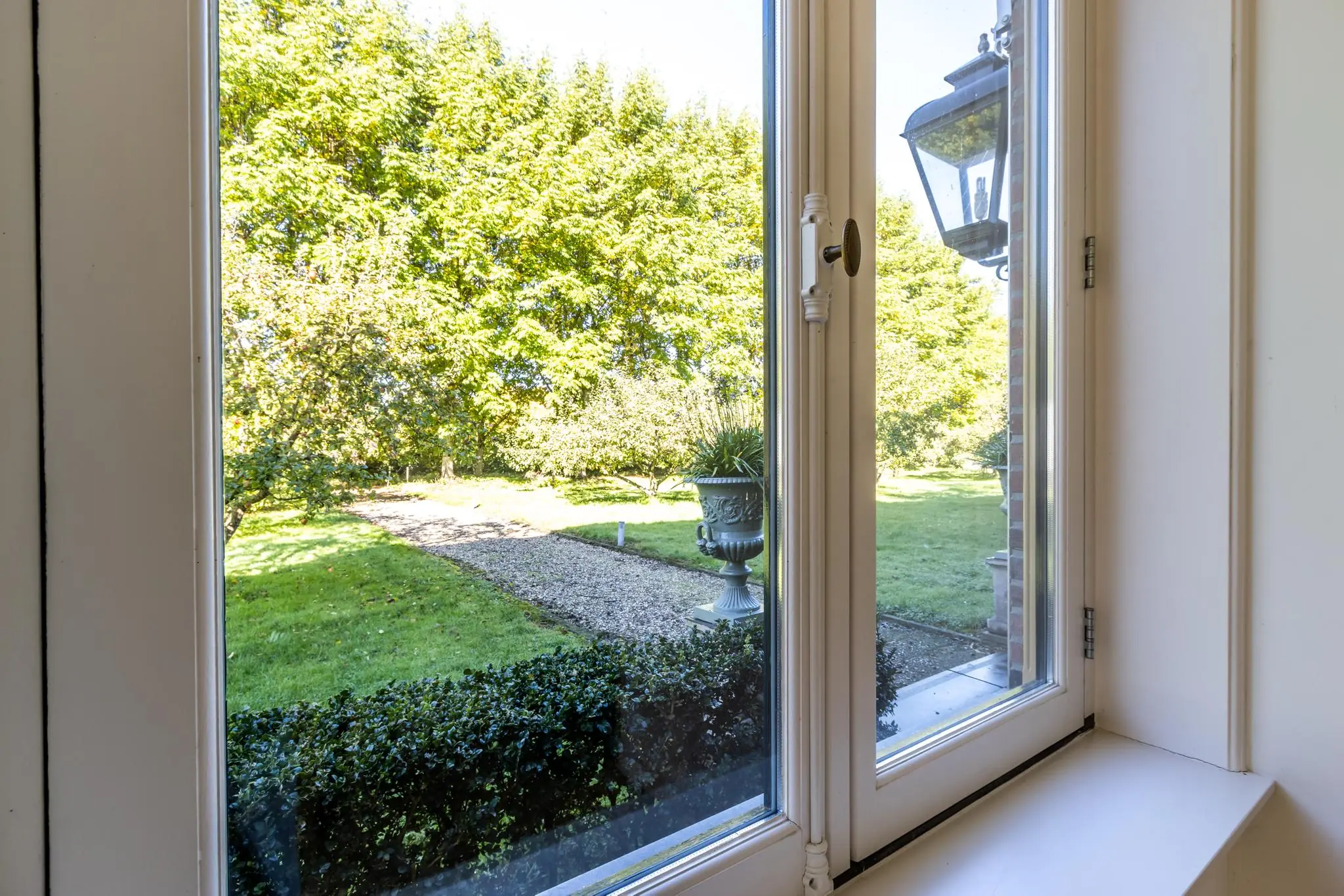 Uitzicht vanuit een raam op een goed onderhouden tuin met grindpad, buxushaag en grote plantenpot.